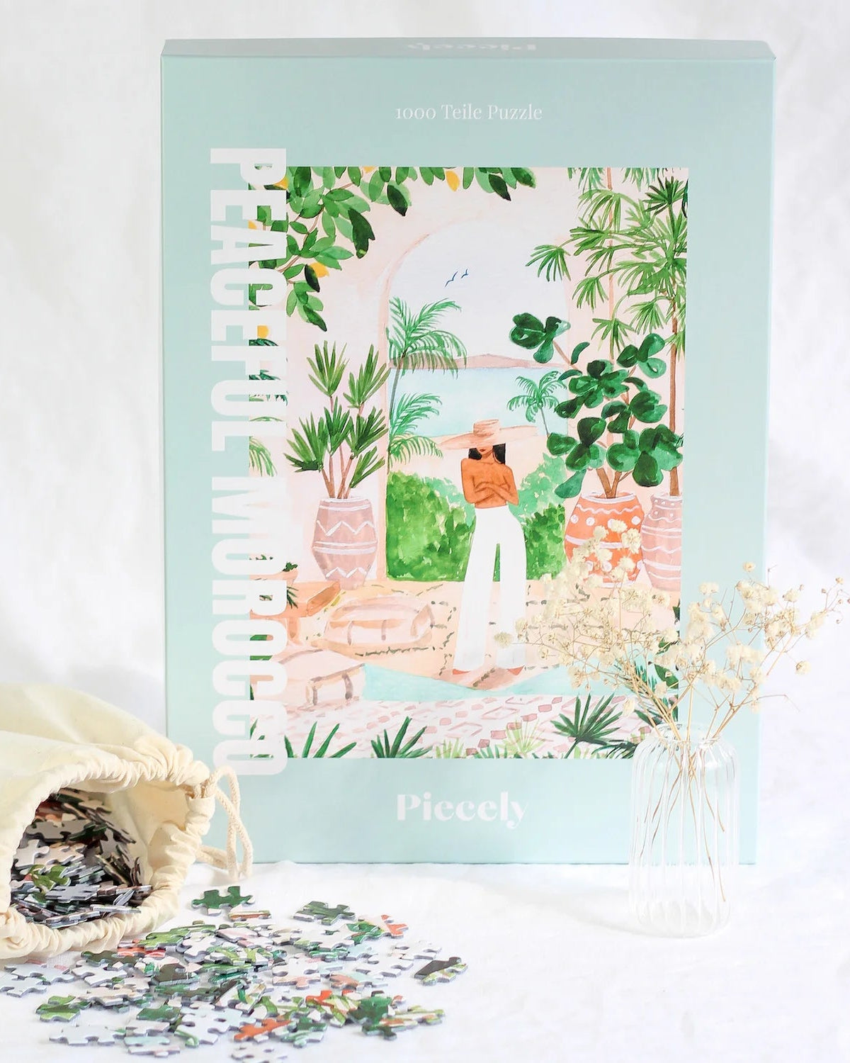 Man sieht die Schachtel des Puzzles "Peaceful Morocco". In der Mitte befindet sich eine Frau mit schwarzen Haaren, Sie trägt einen großen Hut, der ihr Gesicht bedeckt. Ihre Arme sind verschränkt, sie trägt kein Oberteil und eine weiße, weite Hose. Um sie herum sind große Töpfe mit Pflanzen. Hinter ihr ist ein Durchbruch und man sieht das Meer und Berge. Vor der Schachtel liegt das Jutesäckchen mit den Puzzleteilen.