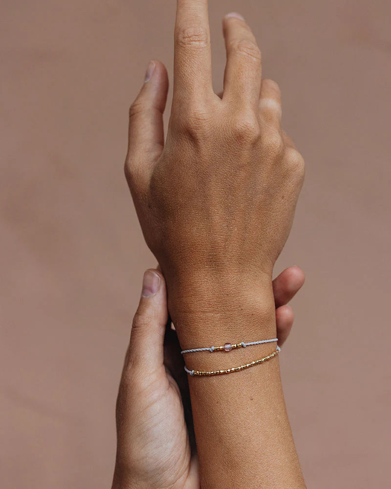 Auf dem Bild sieht man eine Hand mit Handgelenk mit einem Armband aus Perlen und einer Glasperle in Blau. Darunter ist ein zweites Armband aus goldenen kleinen Perlen.