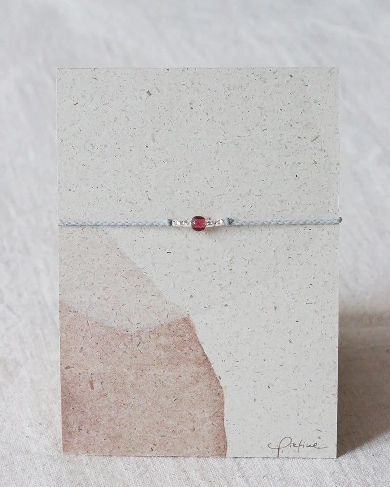 Auf dem Bild sieht man eine Graspapier Pappe mit einem Armband aus Perlen und einer Glasperle in Beere.