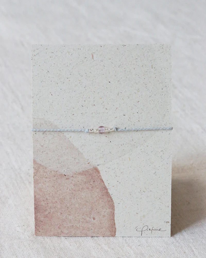 Auf dem Bild sieht man eine Graspapier Pappe mit einem Armband aus Perlen und einer Glasperle in Malve.