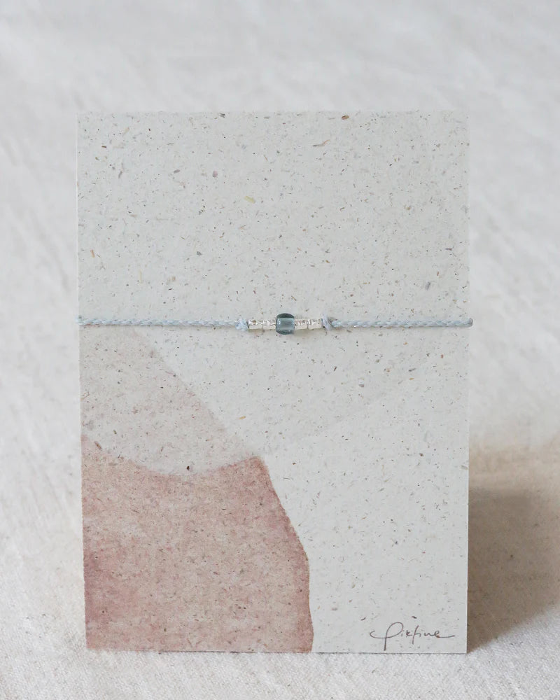 Auf dem Bild sieht man eine Graspapier Pappe mit einem Armband aus Perlen und einer Glasperle in Tannenblau.