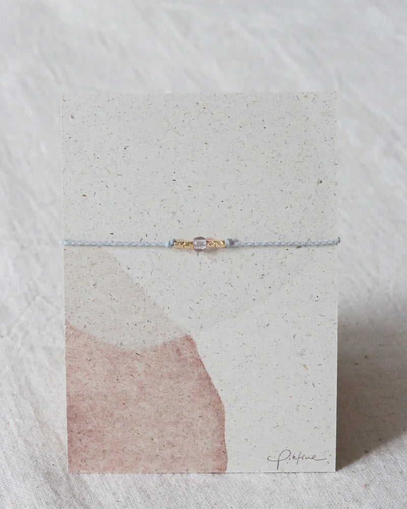 Auf dem Bild sieht man eine Graspapier Pappe mit einem Armband aus Perlen und einer Glasperle in Malve.