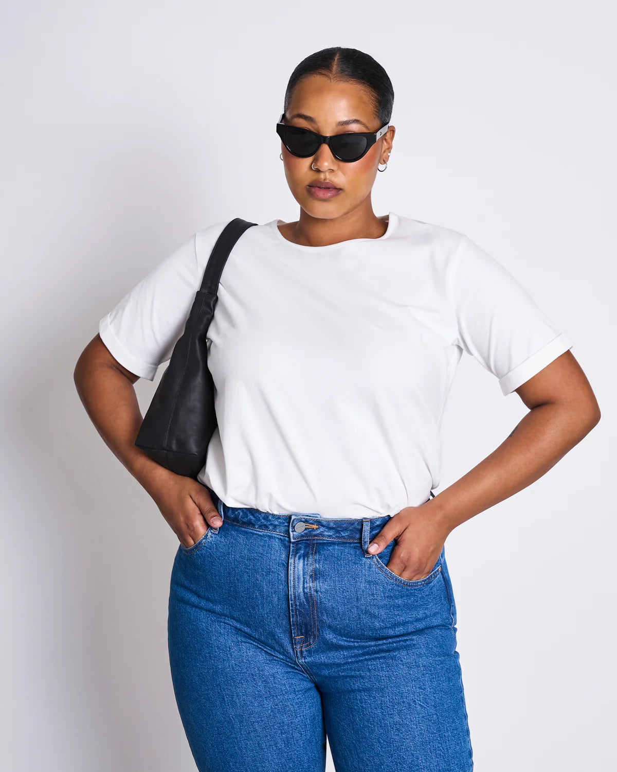 Eine Frau mit dunkler Sonnenbrille und zurückgebundenen Haaren trägt ein weißes T-Shirt mit geradem Schnitt und eine hochgeschnittene blaue Jeans. Sie hat eine schwarze Tasche über die Schulter gelegt und posiert mit den Händen in den Hüften vor einem weißen Hintergrund.
