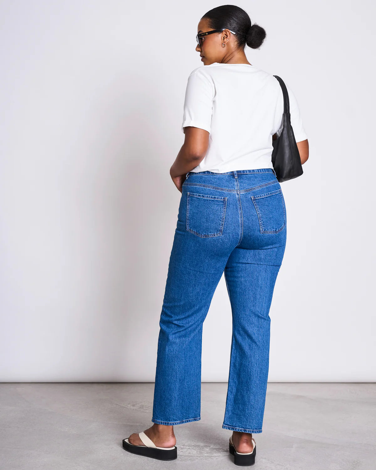 Rückansicht einer Frau mit schwarzer Tasche über der Schulter. Sie trägt ein weißes T-Shirt, eine blaue Jeans und offene Sandalen. Der schlichte, weiße Hintergrund hebt das Outfit hervor.