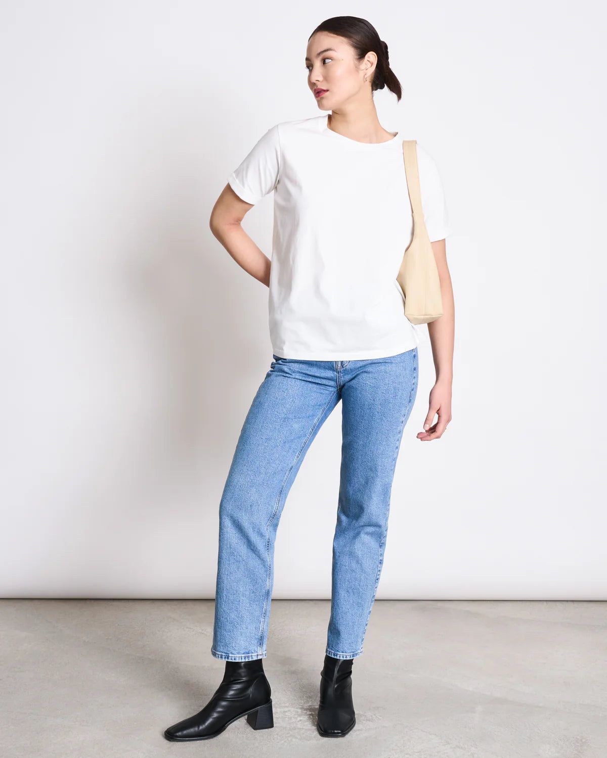 Eine Frau steht entspannt vor einem weißen Hintergrund. Sie trägt ein weißes T-Shirt, eine blaue Jeans und schwarze Stiefeletten. Eine beigefarbene Stofftasche hängt locker über ihrer Schulter.