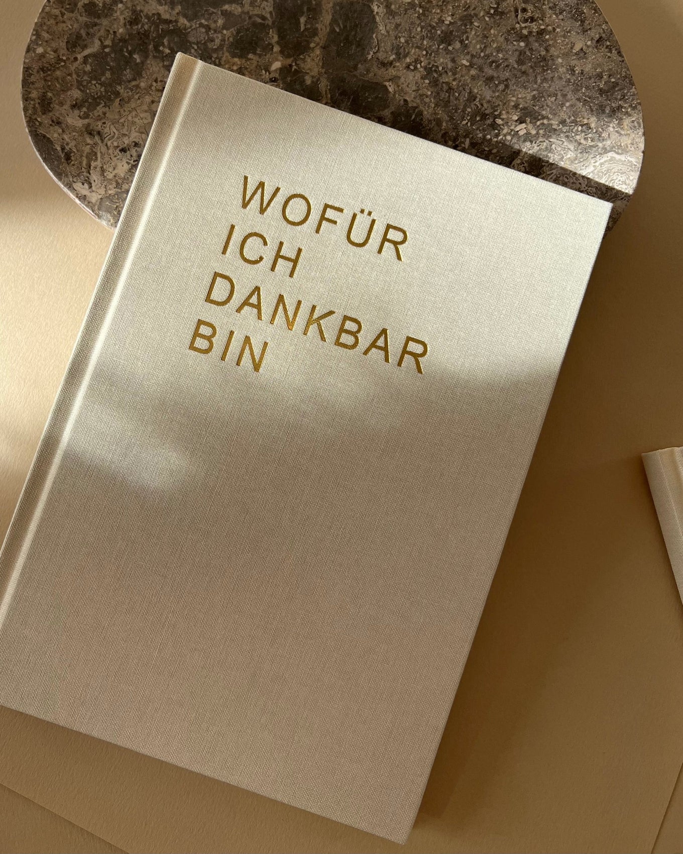 Auf dem Bild sieht man das Dankbarkeitstagebuch, wie es im oberen Bereich auf einem Mamorstein liegt. Es ist in beigen Leinenstoff eingeschlagen und in einer goldenen Prägung steht auf der Vorderseite "Wofür ich dankbar bin". 