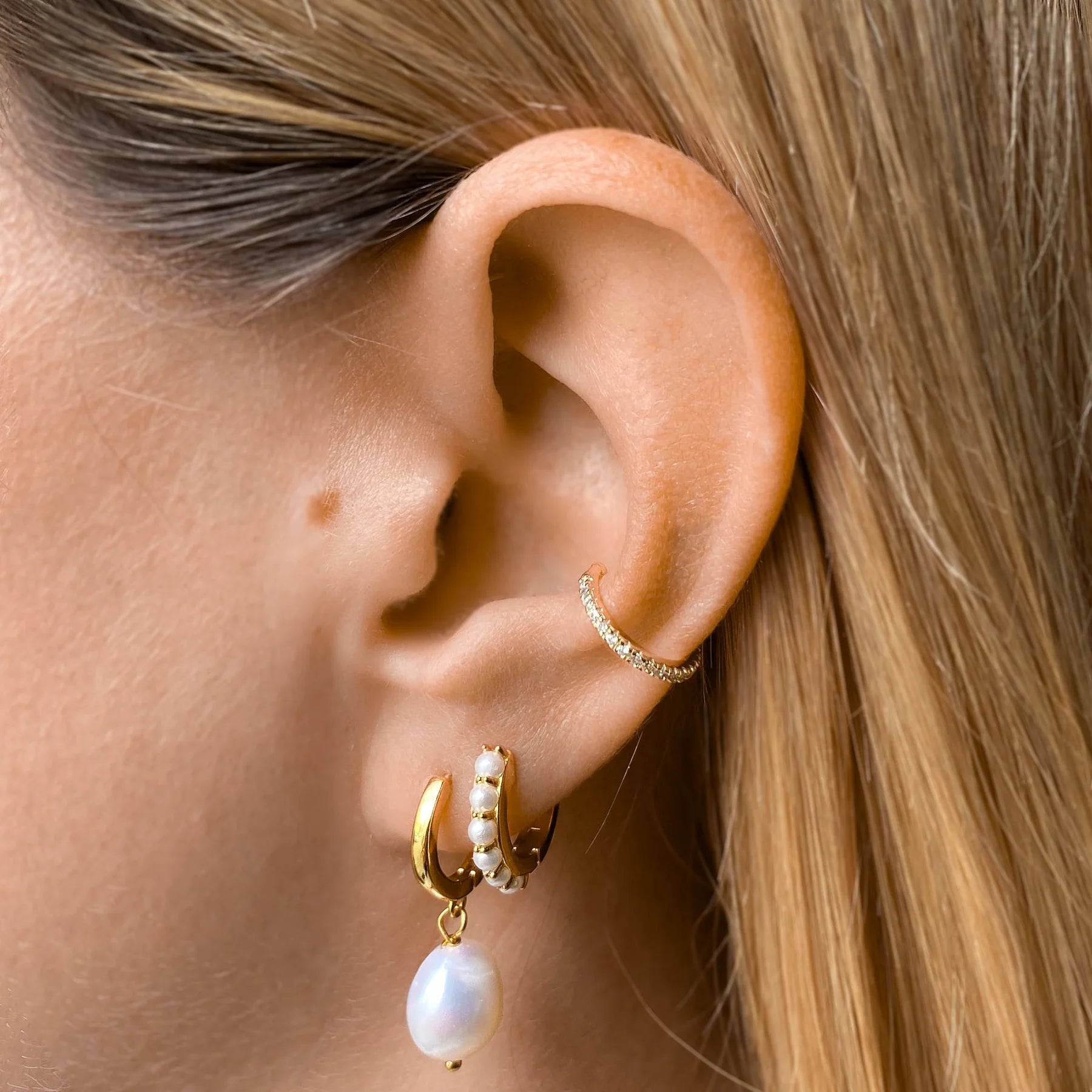 Nahaufnahme eines Ohrs mit fair produzierten goldenen Creolen, einem Perlenanhänger-Ohrring und einem funkelnden Earcuff in Gold. 