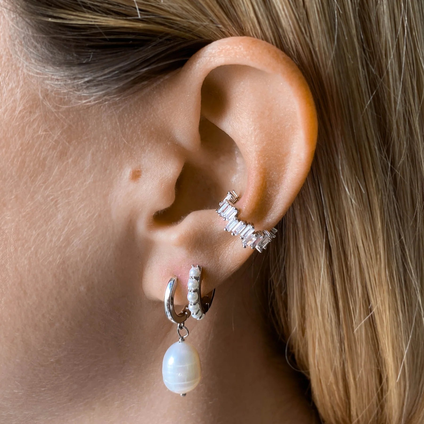 Nahaufnahme eines Ohrs mit fair produzierten silbernen Creolen, einem nchhaltigen Perlenanhänger-Ohrring und einem funkelnden Earcuff. 