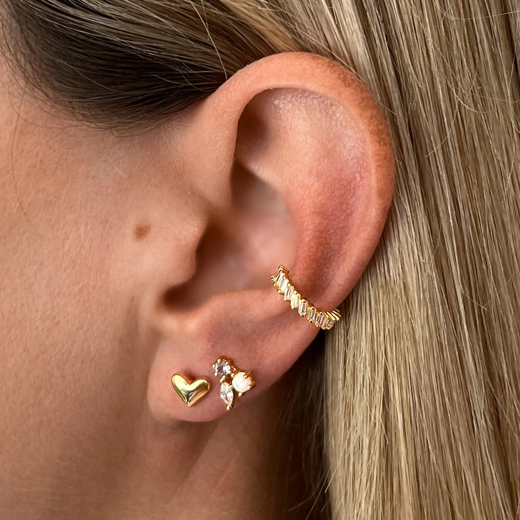 Nahaufnahme eines Ohrs mit einem nachhaltigen goldenen Herz-Ohrstecker, einem funkelnden Ohrstecker und einem fair produzierten Earcuff.