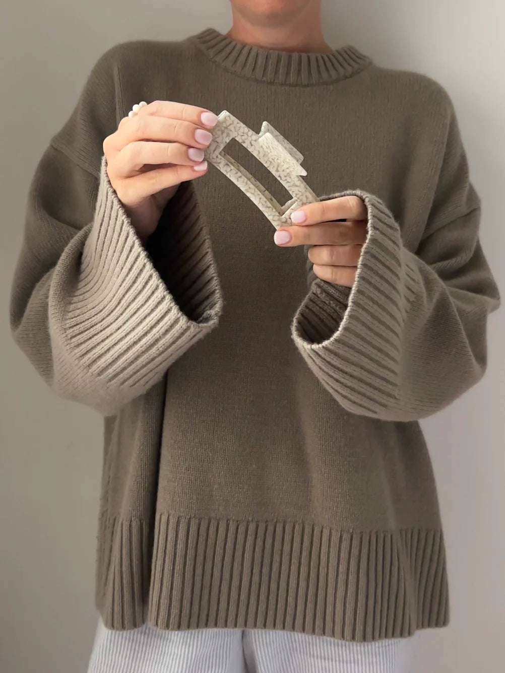 Oberkörper einer Frau in einem khaki Pullover, die eine Haarklammer in cremeweiß gesprenkelt aus cellulose acetat in die Kamera hält. 