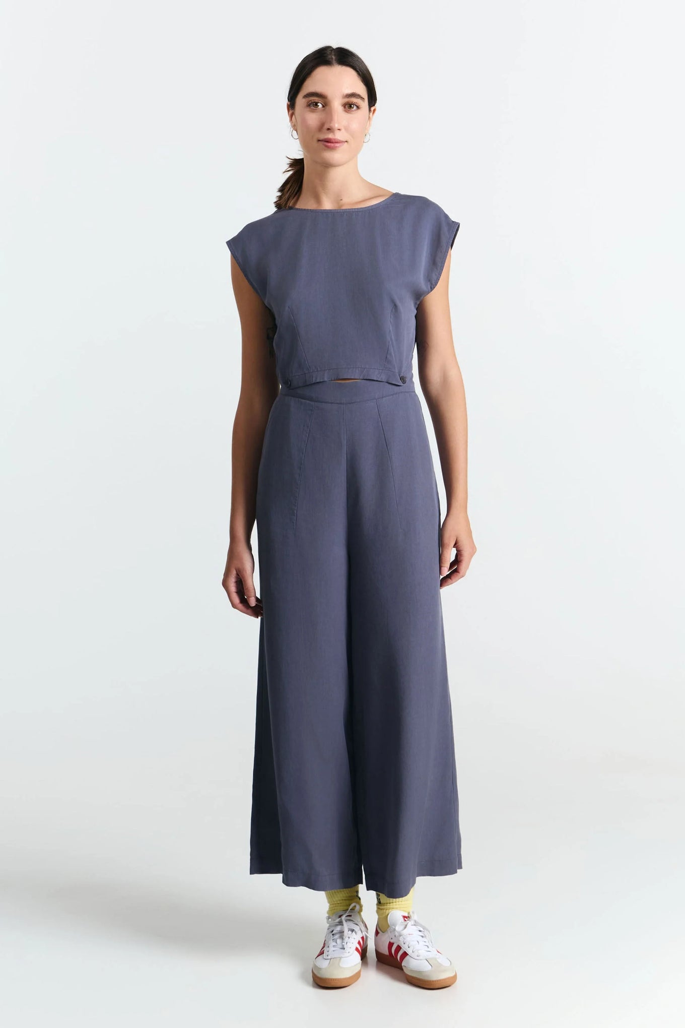 Frau von vorne trägt eine nachhaltige graue Bluse und dazu eine fair produzierte weite Hose in grau. Durch Knöpfe werden beide Teile zum Jumpsuit. 