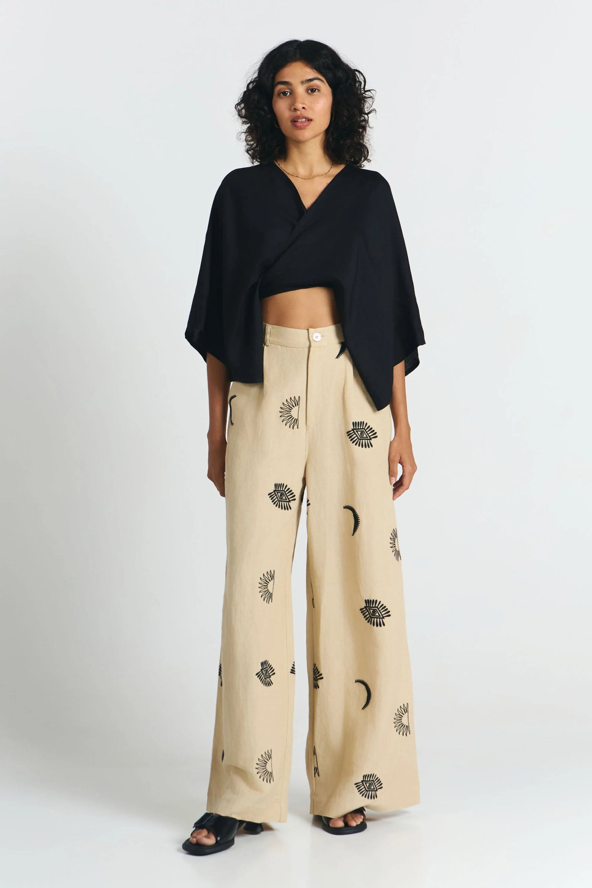 Frau von vorne trägt eine nachhaltige schwarze Bluse und dazu eine fair produzierte weite Hose in beige mit Stickereien aus Mond, Sonne, Augen. 
