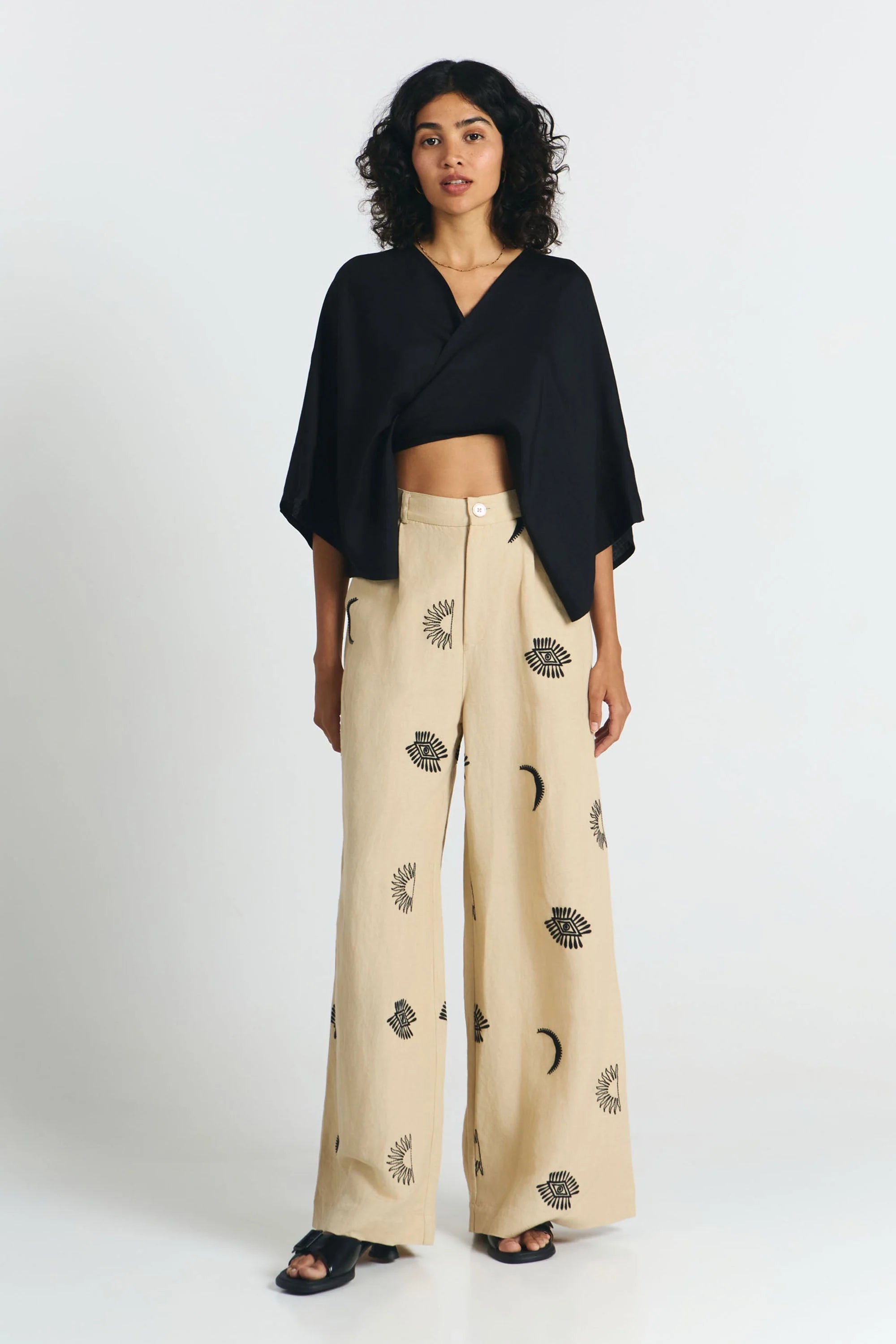Frau von vorne trägt eine nachhaltige schwarze Bluse und dazu eine fair produzierte weite Hose in beige mit Stickereien aus Mond, Sonne, Augen. 
