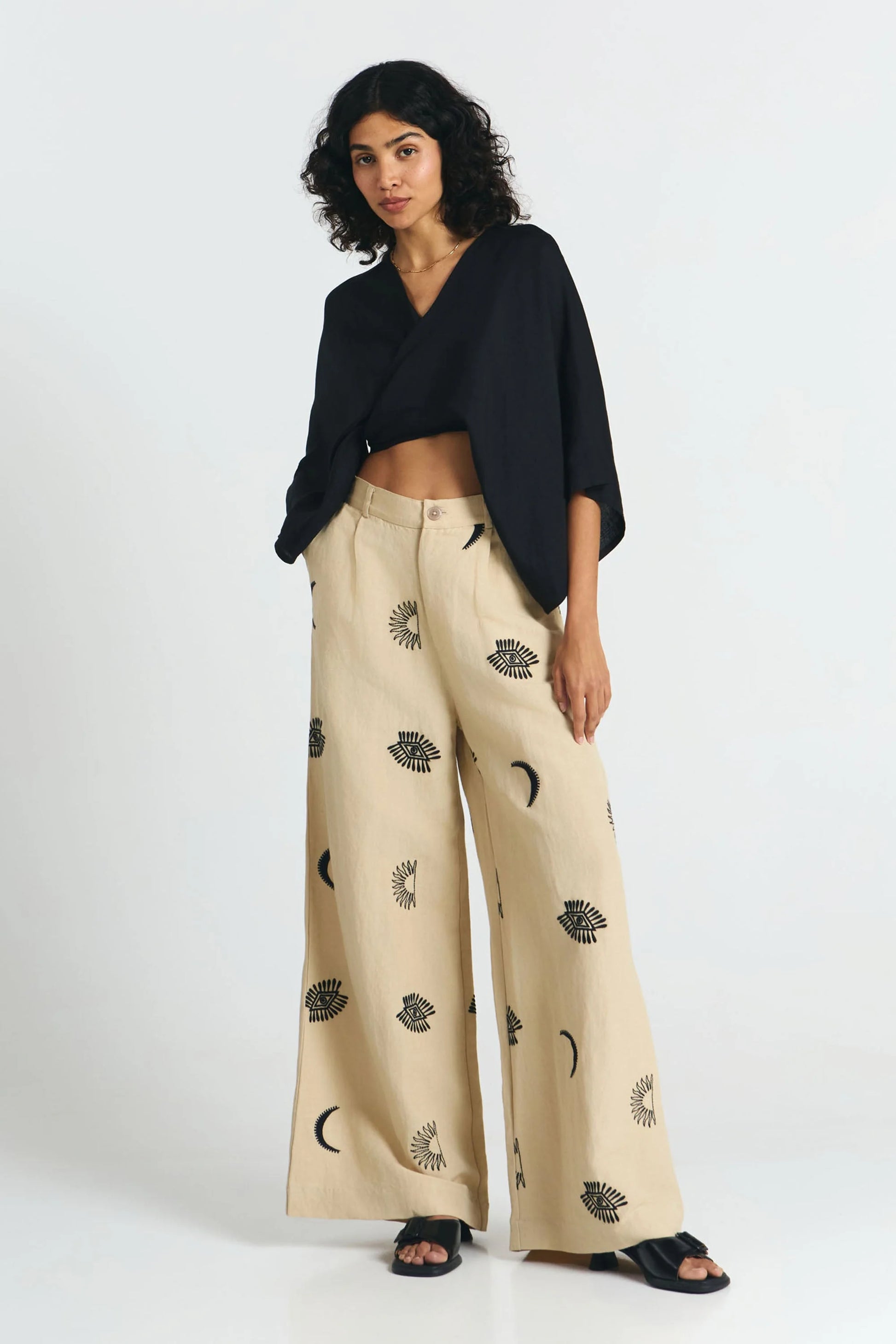 Frau von vorne trägt eine nachhaltige schwarze Bluse und dazu eine fair produzierte weite Hose in beige mit Stickereien aus Mond, Sonne, Augen. 