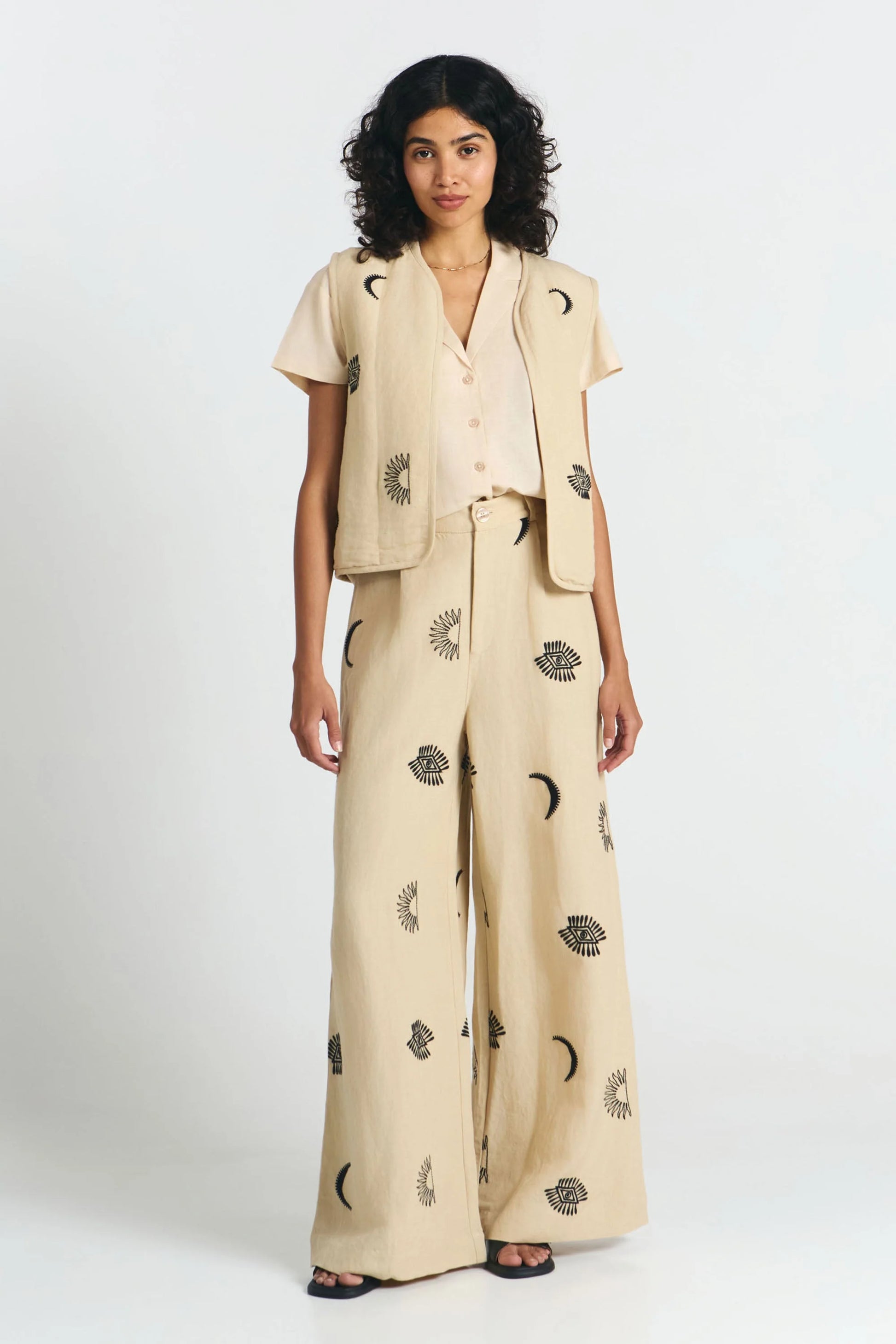 Frau von vorne trägt eine nachhaltige beige Bluse und dazu eine fair produzierte Weste in beige mit Stickereien aus Mond, Sonne, Augen und die passende weite Hose in beige mit Stickereien aus Mond, Sonne, Augen. 