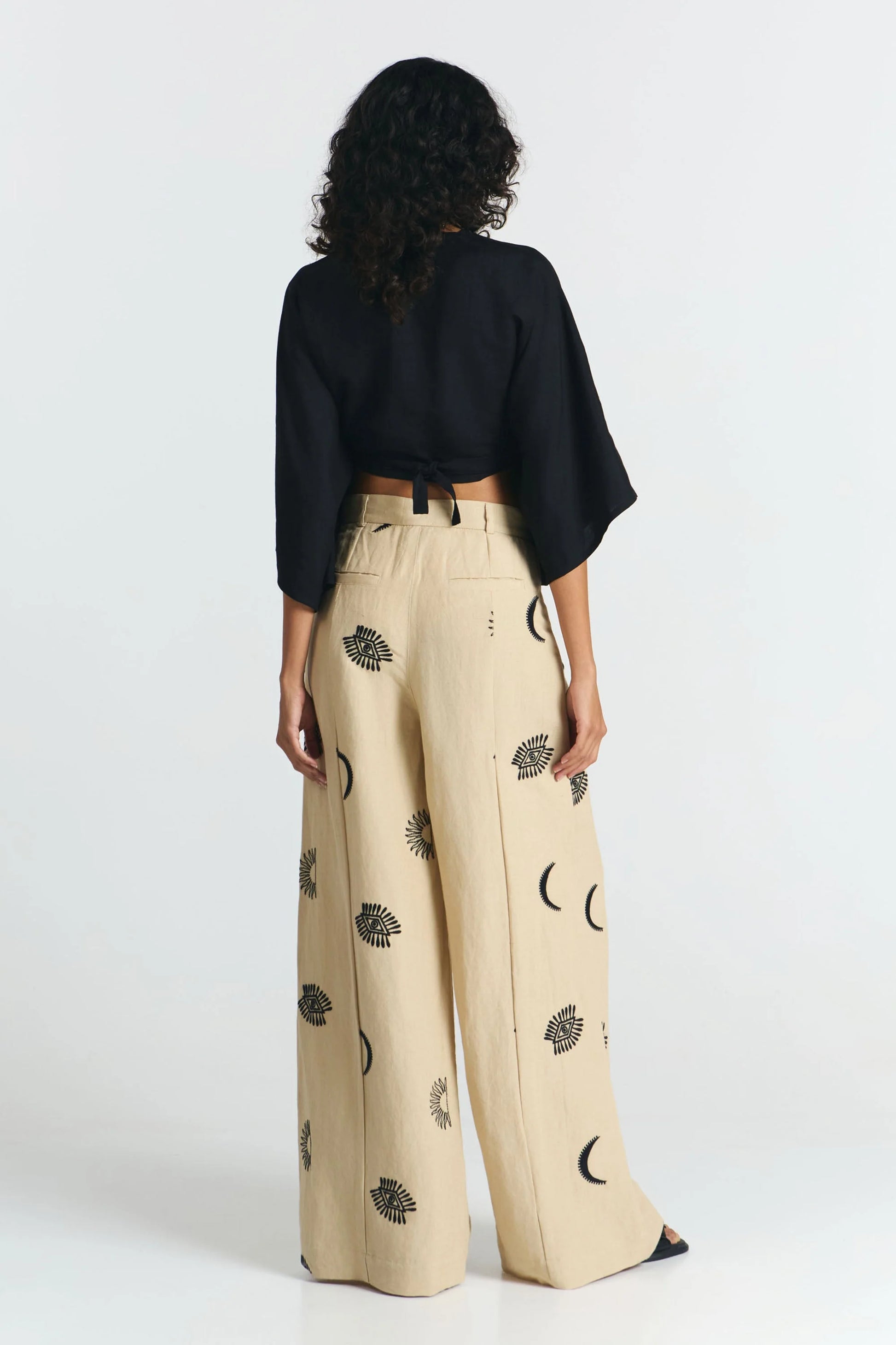Frau von hinten trägt eine nachhaltige schwarze Bluse und dazu eine fair produzierte weite Hose in beige mit Stickereien aus Mond, Sonne, Augen. 